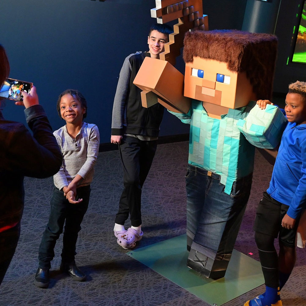 Over the shoulder photo of woman using her camera to take a photo of three older children posing with a life sized sculpture of Steve from Minecraft. 